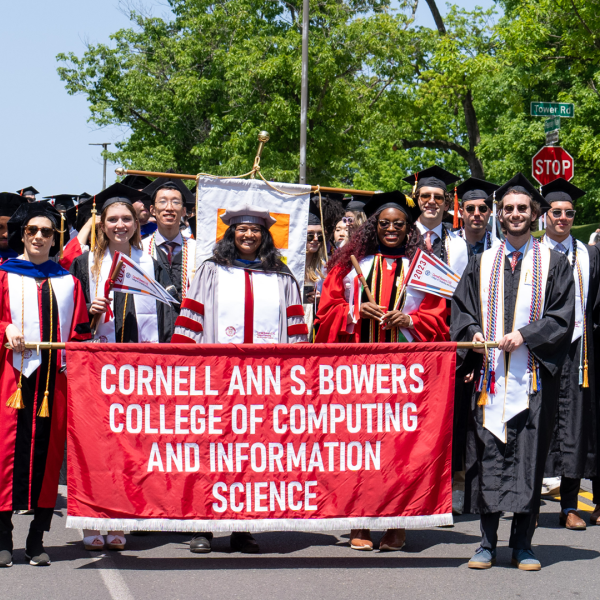 Several students in graduation robes march down a street on sunny day holding a banner that says Cornell Ann S. Bowers College of Computing and Information Science.