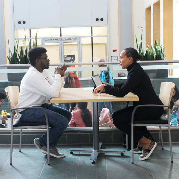 A color photo of 2 people sitting at a table talking