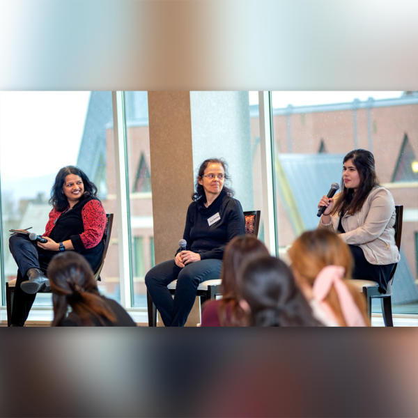 A color photo of 3 women sitting on a stage. One of them speaking into a microphone