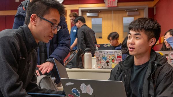 A color photo showing 2 students speaking near a laptop