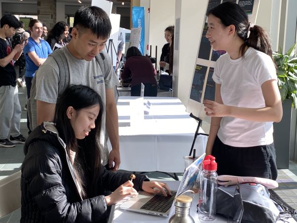 A color photo showing two people working on a laptop computer with another person looking on